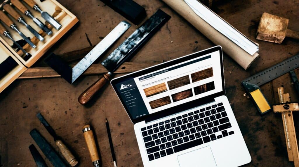Laptop mit einer geöffneten Handwerker-Website steht zwischen Stechbeiteln, Säge und Zollstock auf einer Werkbank